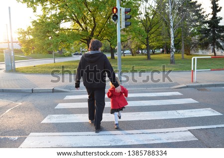 A man and a small child on a zebra crossing trespassing by crossing  the street on red flashing lights. Father is wearing black clothes, the girl has a red coat and striped stockings. Behind view.