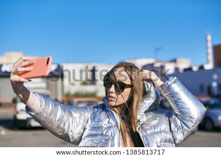 Beautiful girl makes selfies on the phone while standing parked outdoors. Dressed in stylish clothes, sunglasses