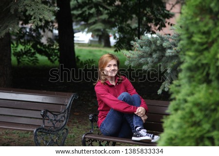 A young girl in casual style in burgundy sweater, jeans and sneakers sits on a bench in a summer park. Around trees and other greens.