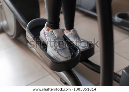 Female legs in fashionable white sneakers stand walking on a stepper simulator in a fitness studio. Young woman trains in a modern gym. Concept of sports and health. Close-up.