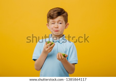 little boy try to taste a fresh lime, sour taste, make grimace, facial emotions negative, in blue T-shirt, isolated yellow background, copy space Royalty-Free Stock Photo #1385864117