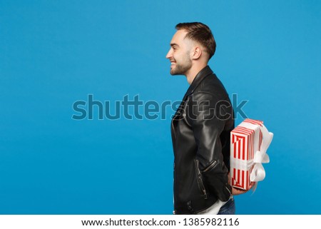 Handsome stylish young unshaven man in black leather jacket white t-shirt hold gift box isolated on blue wall background studio portrait. People sincere emotions lifestyle concept. Mock up copy space