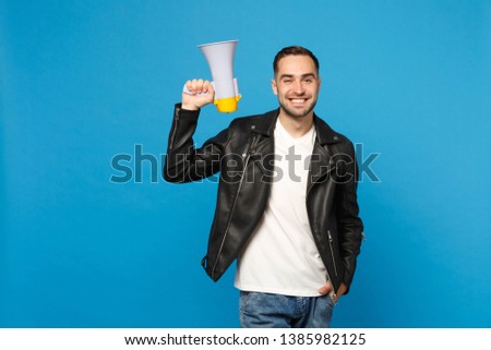 Handsome young unshaven man in black jacket white t-shirt scream in megaphone, announces discounts sale isolated on blue wall background studio portrait. People lifestyle concept. Mock up copy space