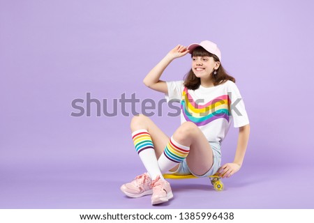 Pretty joyful teen girl in vivid clothes sitting on yellow skateboard, looking aside isolated on violet pastel wall background in studio. People sincere emotions lifestyle concept. Mock up copy space