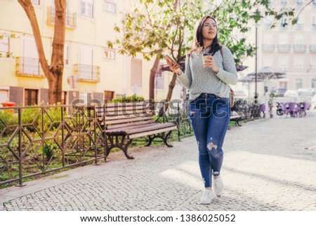 Content woman walking in city with smartphone and plastic cup. Pretty young lady going along walkway with trees and buildings in background. City walk concept. Full length front view.