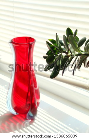 Red vase on a white window sill next to a flower indoor and a shadow from the window blinds, selective focus.