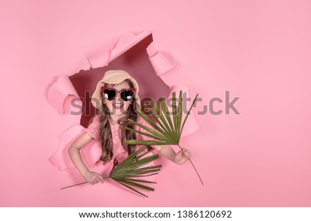 funny little girl looks out from the hole in a beach hat and glasses in the shape of a heart, in the hands of tropical leaves, on a colored background, a place for text, Studio shooting