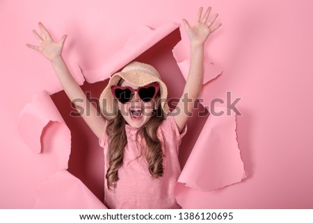 funny little girl peeking out of a hole in a beach hat and heart-shaped glasses on a colored background, place for text, Studio shooting
