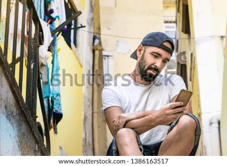Portrait of a spanish man with beard, casual style, sending messages - chatting with his smartphone mobile, sitting on the street, outdoors.