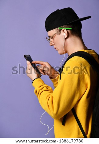 Portrait of a joyful young student in a yellow sweater listening to music with headphones and selects the track song on the smartphone screen isolated over a purple background