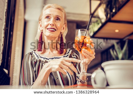 Fashionable short-haired woman. Elegant short-haired old woman wearing striped blouse and gently holding glass with refreshing drink