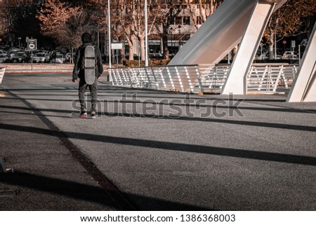 Young teenage skater walking on an industrial footbridge in Rome, Italy, called " Ponte della Musica". Blurry urban background. Tactical back pack with board strappen on. Grey and black color scheme.