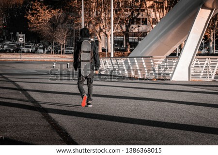 Young teenage skater walking on an industrial footbridge in Rome, Italy. Extreme action sports concept, golden hour, natural light, blurry urban background. Tactical back pack with board strappen on.
