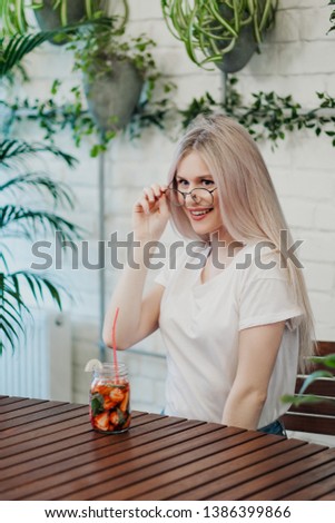 Young beautiful athletic woman blonde in glasses sits at a table in a green cafe and drinks strawberry lemonade from a glass jar. Smiles and laughs.