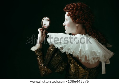 Who's that pretty. Medieval redhead young woman in golden vintage clothing as a duchess looking in the mirror on dark green background. Concept of comparison of eras, modernity and renaissance.