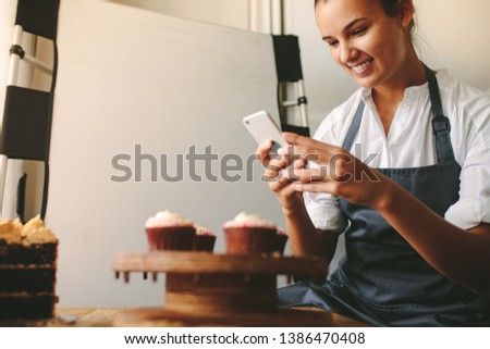 Young woman taking a pictures while cooking tasty pastry in the kitchen. Happy woman taking a picture of cupcakes on a wooden board with mobile phone while standing at the kitchen.