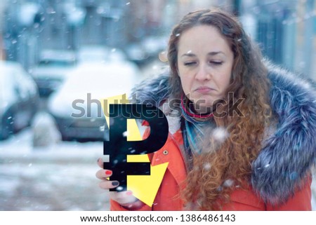 Exaggeratedly sad red-haired girl in warm clothes in winter with a ruble sign and an arrow down. Concept - the ruble falls