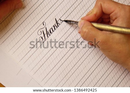 female hand writes with the inky pen the word thanks on a white paper sheet with stripes. stationery on desk close up top view. spelling lessons and caligraphy exercises. Template, layout, background.