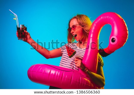 Female half-length portrait on blue neon lights studio background. Woman in red sunglasses and rubber ring as a flamingo with drink and candy. Facial expression, summer, weekend concept. Trendy colors