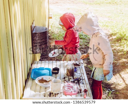 Two young girls sisters play outdoors in so called mud kitchen, where you can make fake food, play with sand, dirt, water, plants and make a mess, it develops imagination and exploration. Royalty-Free Stock Photo #1386539579