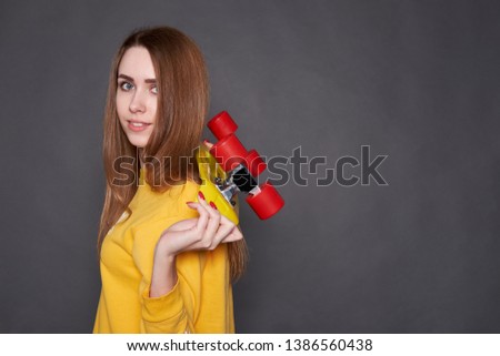 Redhead attractive girl wit long hair wearing yellow sweatshirt holding yellow skateboard with red wheels. Studio shot on grey background. Active lifestyle. Copy space. 