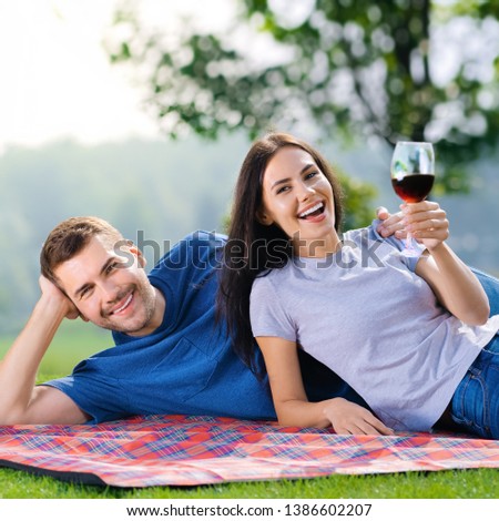 Bright photo of young happy couple in love, lying together on a picnic blanket, outdoors, at sunny day. Making a toast with red wine glass. Square compostion picture.