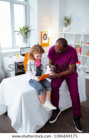Cartoon and pediatrician. Cute little girl feeling entertained while watching cartoon on tablet while visiting pediatrician