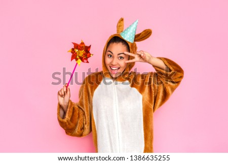 Young woman in bunny kigurumi wearing party hat standing isolated on pink background holding pinwheel posing to camera gesturing cool smiling happy