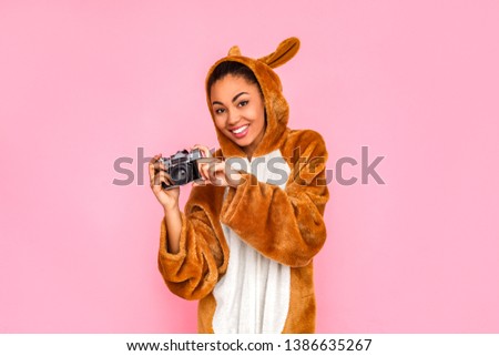 Young woman in bunny kigurumi standing isolated on pink background holdign film camera taking pictures smiling cheerful