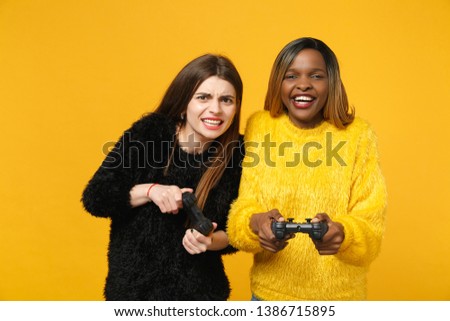 Two young women friends european and african american in black yellow clothes hold joystick isolated on bright orange wall background, studio portrait. People lifestyle concept. Mock up copy space.