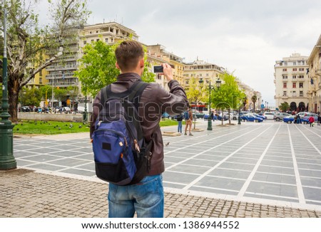 Young tourist with backpack taking pictures of a square Archeas Agoras and a pedestrian street on a smartphone in Thessaloniki, Greece in spring.