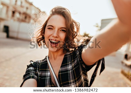 Wonderful curly woman with dark eyes playfully posing on the street. Outdoor photo of inspired young lady making selfie.