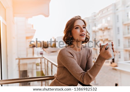 Dreamy dark-eyed girl drinking tea at balcony. Photo of caucasian well-dressed female model holding cup of coffee.