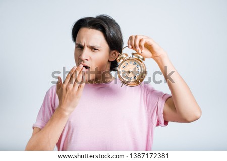 handsome young man yawns with alarm clock, hipster, studio photo on gray background