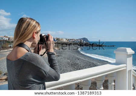 Young millennial female of Caucasian ethnicity, a remote photographer or a solo tourist, holding a digital, professional camera and taking pictures of the ocean and the small pebble beach behind