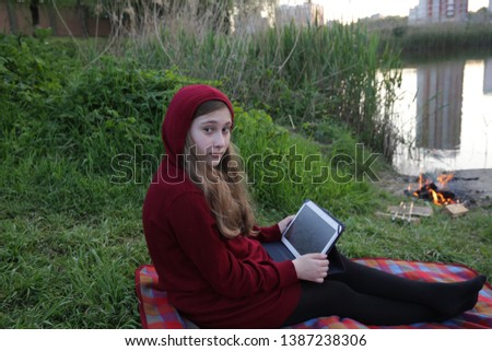 teenager girl with long hair in red hoodie uses tablet sitting on the green grass by the fire with lake view and reflection of urban buildings in the lake