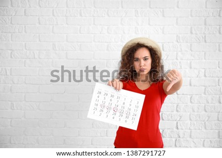 African-American woman holding calendar with marked days of menstruation and showing thumb-down on white background