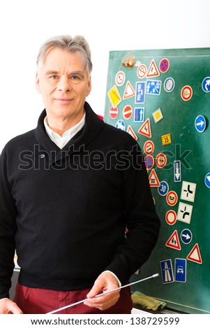 Driving school - driving instructor in his class, he standing in front of a blackboard and looking in the camera, in the background are traffic signs