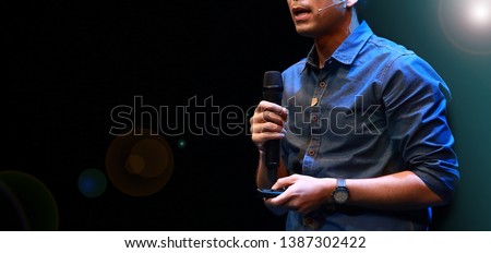 The male speaker is doing the public speaking with the dark background under the spot light and lense flare, in concept of talk show, flare light, motivational speaking, inspiration speaker. Royalty-Free Stock Photo #1387302422