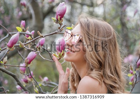 Young blonde woman near blossoming magnolia flowers tree in spring park on sunny day. Magnolia trees. Beautiful happy girl enjoying smell in a flowering spring garden