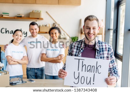 selective focus of handsome man holding placard with thank you text while standing near multicultural volunteers in charity center