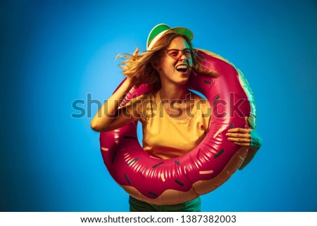 Beautiful female half-length portrait isolated on blue neon lights studio background. Woman in green cap, rubber ring and sunglasses. Facial expression, summer, weekend concept. Trendy colors.