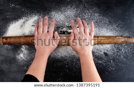 Brown wooden rolling pin in female hands on a black background with white wheat flour, top view