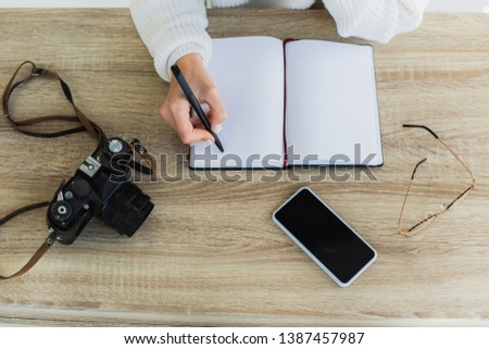 cropped view of young woman holding pen near empty notebook, digital camera, glasses and smartphone with blank screen 