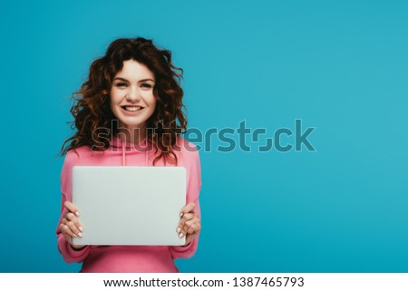 cheerful curly redhead girl holding laptop while standing on blue 