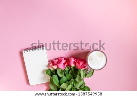 The cup of hot coffee for breakfast, empty notepad with funny pen and bouquet of fresh roses on soft pink background.