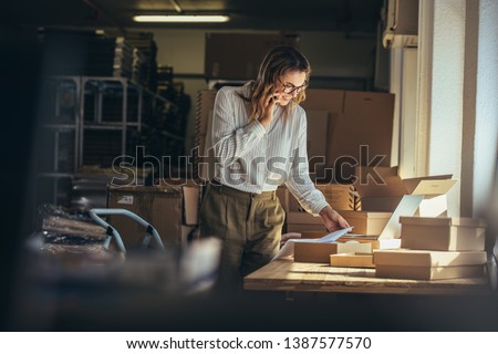 Woman online seller confirming orders from customer on the phone. E-commerce business owner looking at the papers and talking on phone in store warehouse. Royalty-Free Stock Photo #1387577570