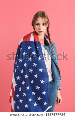 Young pretty girl with two braids in denim jacket with headphones and big american flag on shoulder dreamily looking in camera over pink background