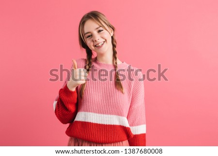 Young cheerful girl with two braids in sweater showing thumb up gesture joyfully looking in camera over pink background