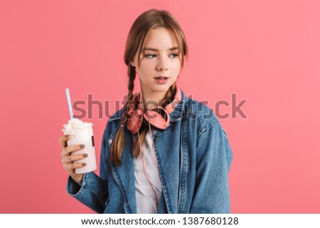 Young attractive girl with two braids in denim jacket with headphones holding milkshake in hand dreamily looking aside over pink background